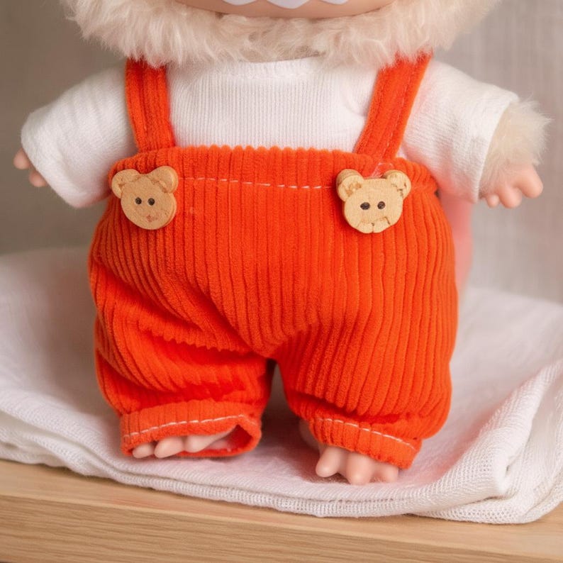 Peut inclure: Une poup&eacute;e v&ecirc;tue d'une salopette en velours c&ocirc;tel&eacute; orange avec des boutons en bois en forme d'ours et un t-shirt blanc. La salopette a des bretelles et arrive aux genoux. La poup&eacute;e a une t&ecirc;te poilue et de petites mains et pieds.