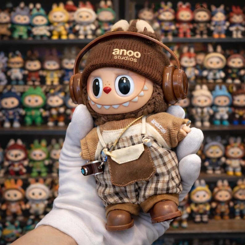 May include: A brown and beige Anoo Studios figure with headphones, a camera, and plaid overalls. The figure has a large head, big eyes, and a toothy grin. The figure is held in a gloved hand, with a shelf of similar figures in the background.