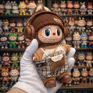 May include: A brown and tan collectible vinyl figure of a cartoon character wearing a knit hat with headphones. The figure has a smiling face, a camera, and plaid overalls. The figure is held in a gloved hand, with other figures in the background.