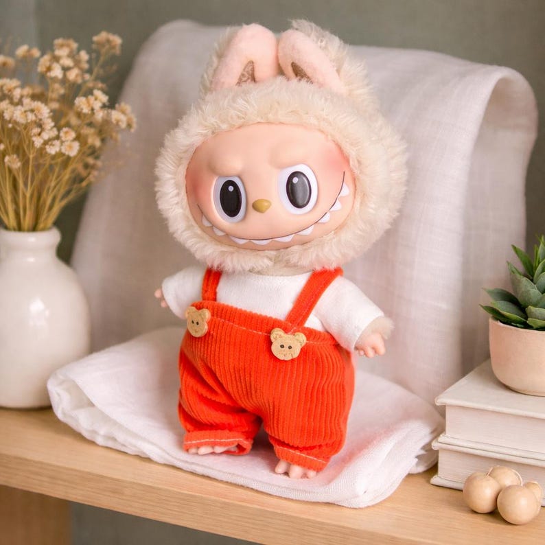 Peut inclure: Une poup&eacute;e en peluche au design unique. La poup&eacute;e pr&eacute;sente un visage souriant avec de grands yeux, portant une capuche en fourrure avec des oreilles de lapin et une salopette en velours c&ocirc;tel&eacute; orange. La salopette a des boutons en forme d'ours. La poup&eacute;e est pos&eacute;e sur un tissu blanc.