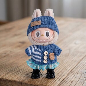 Op de afbeelding: Een grillige pop met een blauwe gebreide muts met konijnenoren en een bijpassende trui. De pop heeft een lachend gezicht, een blauw-wit gestreepte trui, een geruite rok en zwarte schoenen. Een kleine patch met een beer staat op de trui.