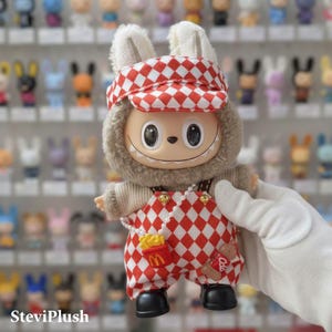K&ouml;nnte beinhalten: Eine Pl&uuml;schtierfigur mit einem hasen&auml;hnlichen Kopf und einem l&auml;chelnden Gesicht. Sie tr&auml;gt einen rot-wei&szlig; karierten Hut und eine Latzhose. Die Latzhose hat eine Pommes-Tasche und ein Coca-Cola-Logo. Die Figur hat schwarze Schuhe und eine Perlenkette.