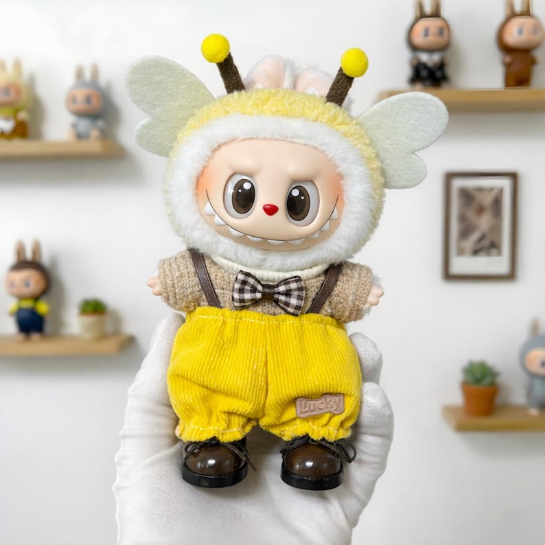 K&ouml;nnte beinhalten: Eine skurrile Puppe im Bienenkost&uuml;m, mit einem gelb-wei&szlig;en Kapuzen-Outfit, Antennen und Fl&uuml;geln. Die Puppe tr&auml;gt einen braunen Pullover, eine gelbe Cordhose und eine karierte Fliege. Die Schuhe der Puppe sind braun.
