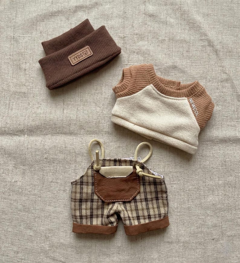 May include: A collection of doll clothes, including a brown headband with a "YYDS" label, a tan and brown sweater with "NEWCOL" text, and plaid overalls with a brown leather pocket.