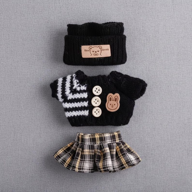 Op de afbeelding: Een zwarte gebreide poppenoutfitset, bestaande uit een muts met een berenlabel, een trui met een konijnenpatch en drie knopen, en een geruite rok. De trui heeft wit-zwart gestreepte mouwen.