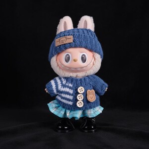 Op de afbeelding: Een grillige pop gekleed in een blauwe gebreide muts met konijnenoren, een bijpassende trui en een geruite rok. De trui heeft een wit en blauw gestreept ontwerp en houten knopen. De pop draagt zwarte schoenen.