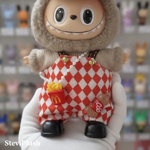 K&ouml;nnte beinhalten: Eine Pl&uuml;schtierfigur mit einem l&auml;chelnden Gesicht und einer pelzigen Kapuze. Sie tr&auml;gt eine rot-wei&szlig; karierte Latzhose mit einer Perlenkette, einer Pommes-Tasche und einer Soda-Dosen-Anstecknadel. Die Figur hat schwarze Schuhe.