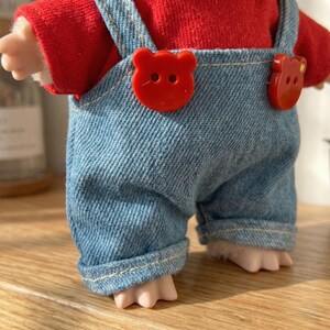 Peut inclure: Une poup&eacute;e v&ecirc;tue d'un haut rouge et d'une salopette en jean bleu. La salopette est orn&eacute;e de boutons rouges en forme d'ours. Les pieds de la poup&eacute;e sont visibles, et le design est mignon et ludique.