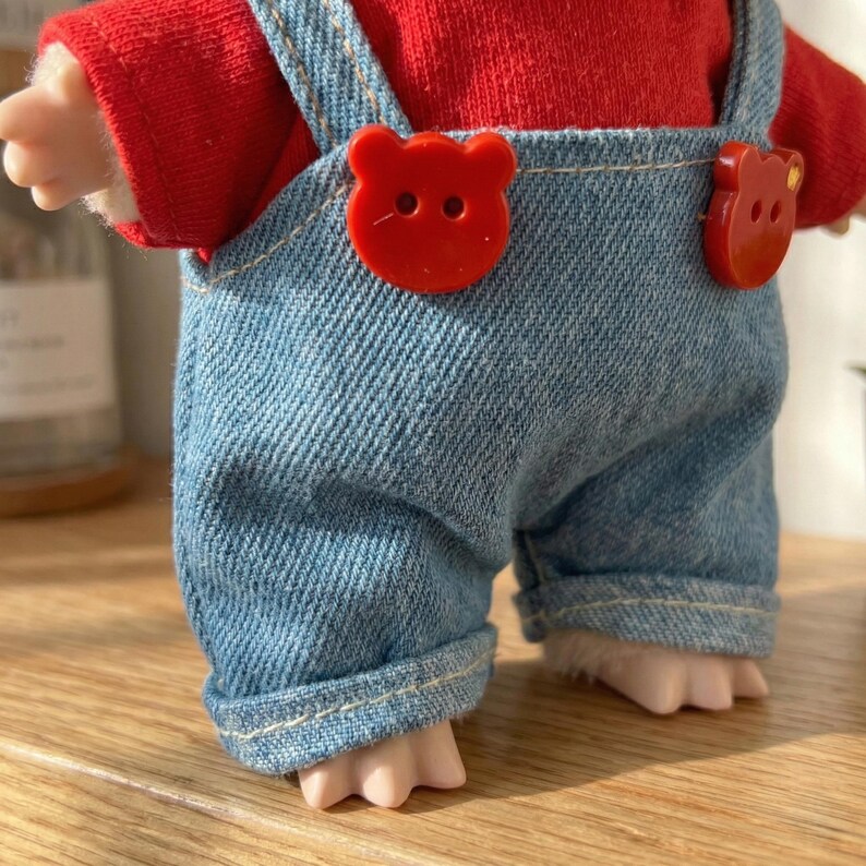 Peut inclure: Une poup&eacute;e v&ecirc;tue d'un haut rouge et d'une salopette en jean bleu. La salopette est orn&eacute;e de boutons rouges en forme d'ours. Les pieds de la poup&eacute;e sont visibles, et le design est mignon et ludique.