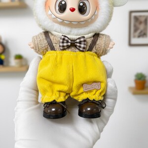 K&ouml;nnte beinhalten: Eine skurrile Puppe mit einem l&auml;chelnden Gesicht, die einen cremefarbenen, pelzigen Hut und einen beigefarbenen Pullover tr&auml;gt. Sie hat gelbe Cordhosen mit dem Wort "Lucky" auf der Tasche, braune Hosentr&auml;ger, eine karierte Fliege und braune Schuhe.