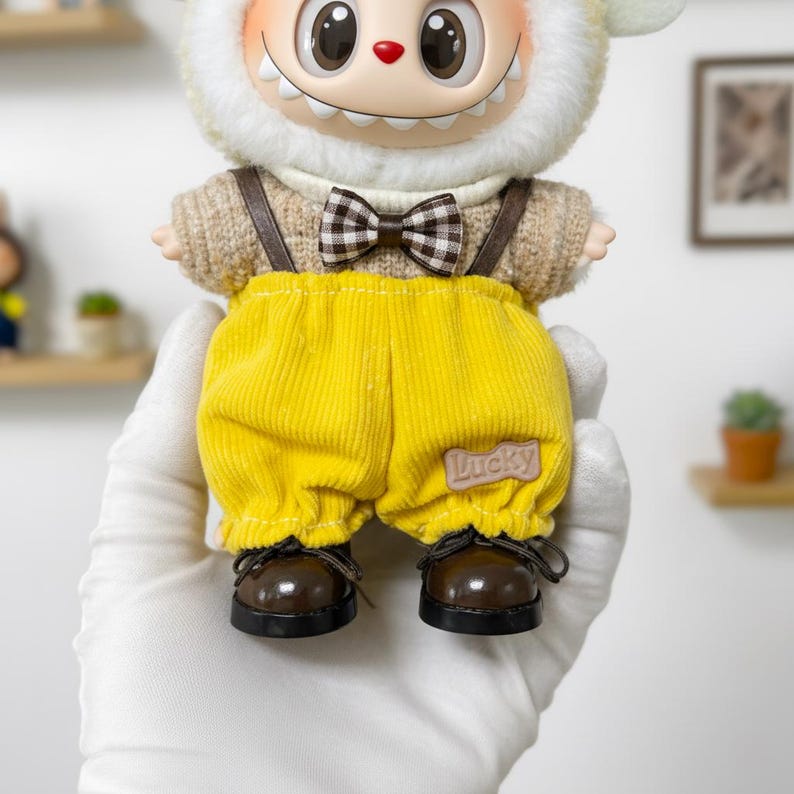 K&ouml;nnte beinhalten: Eine skurrile Puppe mit einem l&auml;chelnden Gesicht, die einen cremefarbenen, pelzigen Hut und einen beigefarbenen Pullover tr&auml;gt. Sie hat gelbe Cordhosen mit dem Wort "Lucky" auf der Tasche, braune Hosentr&auml;ger, eine karierte Fliege und braune Schuhe.