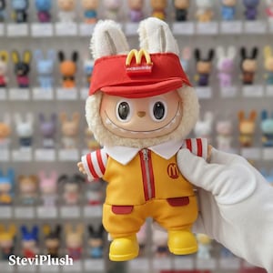 K&ouml;nnte beinhalten: Pl&uuml;schfigur in gelber McDonald's-Uniform mit roten Akzenten. Die Figur hat ein rotes Visier mit dem McDonald's-Logo, einen wei&szlig;en Fellbesatz und Hasenohren. Die Figur wird von einer wei&szlig;en, behandschuhten Hand gehalten.
