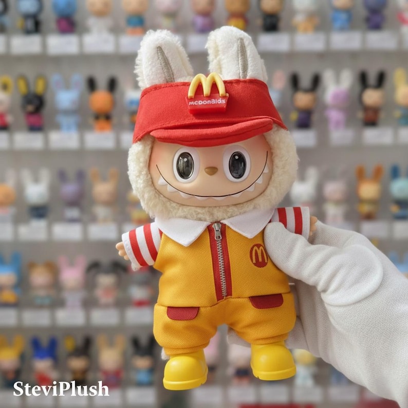 K&ouml;nnte beinhalten: Pl&uuml;schfigur in gelber McDonald's-Uniform mit roten Akzenten. Die Figur hat ein rotes Visier mit dem McDonald's-Logo, einen wei&szlig;en Fellbesatz und Hasenohren. Die Figur wird von einer wei&szlig;en, behandschuhten Hand gehalten.