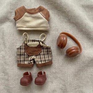 May include: A doll-sized outfit set featuring a brown and cream-colored raglan shirt with the text "NEWCOL", plaid overalls, brown headphones, and two pairs of brown boots. The outfit is designed for a doll.