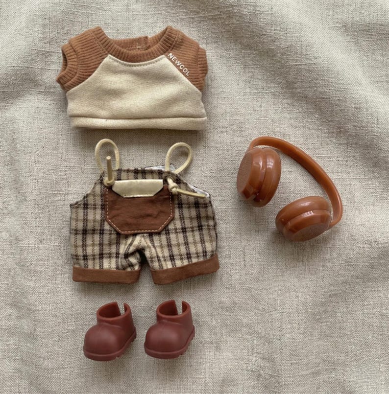 May include: A doll-sized outfit set featuring a brown and cream-colored raglan shirt with the text "NEWCOL", plaid overalls, brown headphones, and two pairs of brown boots. The outfit is designed for a doll.