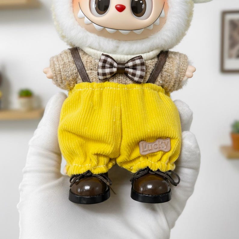 K&ouml;nnte beinhalten: Sammlerpuppe mit l&auml;chelndem Gesicht, tr&auml;gt einen beigen Pullover, gelbe Cord-Latzhose und braune Schuhe. Die Puppe hat eine Fliege und Hosentr&auml;ger. Die Latzhose hat einen "Lucky"-Aufn&auml;her.