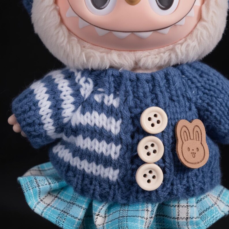 Op de afbeelding: Een close-up van een pop met een blauwe gebreide trui met witte patronen en drie cr&egrave;mekleurige knopen. De pop heeft een witte harige capuchon en een blauwe geruite rok. Een houten konijnvormig embleem is aan de trui bevestigd.