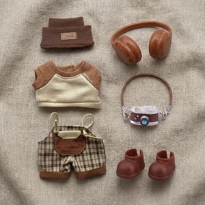 May include: A collection of miniature doll clothing and accessories in shades of brown and beige. Items include a beanie with "YYDS" text, headphones, a shirt with "NEWCOL" text, plaid overalls, a camera, and boots.