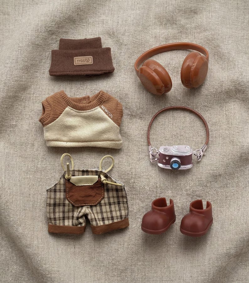 May include: A collection of miniature doll clothing and accessories in shades of brown and beige. Items include a beanie with "YYDS" text, headphones, a shirt with "NEWCOL" text, plaid overalls, a camera, and boots.
