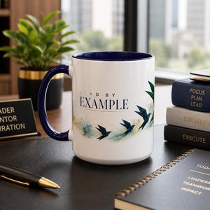 May include: A white ceramic mug with a dark blue interior and handle, featuring the text "LEAD BY EXAMPLE" in dark blue lettering above a graphic of several dark blue origami birds flying amidst light blue and gold watercolor-style accents.