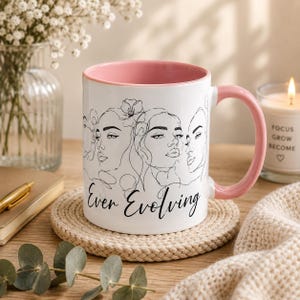 May include: A white ceramic mug with a soft pink interior and handle, featuring black line art illustrations of three faces and the text "Ever Evolving" in a cursive script. The mug has a smooth finish and a cylindrical shape.