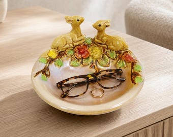 May include: A round ceramic trinket dish featuring two sculpted deer figures resting atop a cluster of colorful painted flowers and green leaves. The dish has a glossy, iridescent finish and contains a pair of tortoise shell glasses and a gold ring.