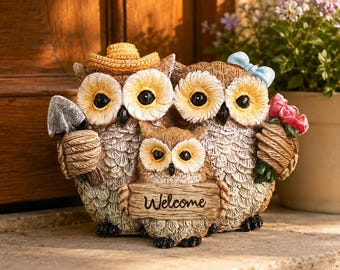 May include: A decorative owl family figurine featuring two adult owls and one owlet. The left adult wears a yellow straw hat and holds a gray trowel. The right adult wears a blue bow and holds pink flowers. The owlet holds a sign reading Welcome.