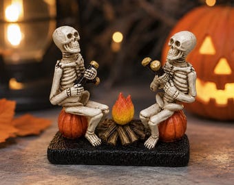 May include: A decorative figurine featuring two off-white skeletons sitting on small orange pumpkins, facing each other around a central campfire with orange and yellow flames, all mounted on a textured black rectangular base.