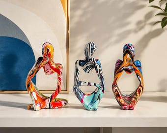 May include: Three abstract human-shaped sculptures in a seated, contemplative pose. The left figure features red, orange, and yellow swirls. The center figure has black, white, and blue patterns. The right figure displays orange, blue, and pink marbled colors.