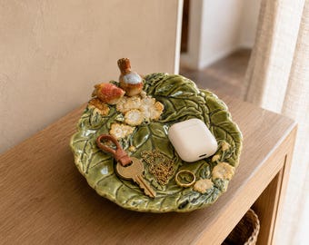 May include: A green ceramic trinket dish shaped like a large leaf with sculpted white flowers and two small birds on the rim. The dish contains a brown leather keychain, a thin gold chain necklace, a gold ring, and a white wireless earbud case.