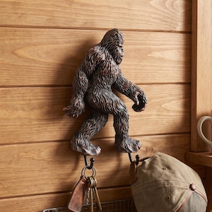Può includere: Un gancio da parete a forma di Bigfoot marrone e testurizzato montato su una superficie in legno. La figura ha due ganci metallici che si estendono dai piedi, reggendo un mazzo di chiavi e un berretto beige. Sotto, un cestino in filo metallico contiene un libro FIELD GUIDE.