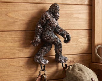 May include: A brown, textured Bigfoot-shaped wall hook mounted on a wooden surface. The figure has two metal hooks extending from its feet, holding a set of keys and a tan baseball cap. Below, a wire basket contains a book labeled FIELD GUIDE.