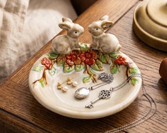 May include: A round ceramic trinket dish featuring two sculpted rabbits sitting atop a decorative rim of painted red berries and green leaves. The dish contains a pair of small pearl earrings, a silver locket necklace, and a silver key pendant.