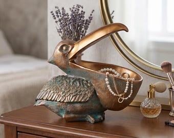 May include: A bronze-toned pelican figurine with a verdigris finish, designed as an open-beak bowl for storage. The pelican features textured feathers and a wide, hollow beak holding a pearl necklace, positioned near a perfume bottle and brushes.