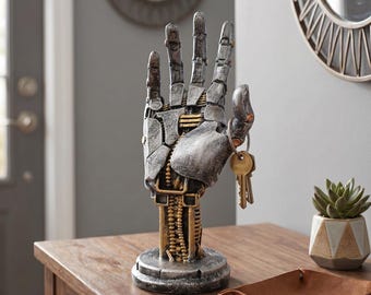 May include: A mechanical hand sculpture featuring a weathered silver and gold finish with segmented plating and exposed internal gear-like details. The upright hand is mounted on a circular base and includes a metal ring on the thumb holding keys.