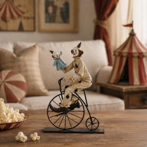 May include: A decorative figurine of a clown riding a black metal penny-farthing bicycle. The clown wears a cream-colored suit with black spade symbols and holds a small blue puppet. The figure is mounted on a rectangular black metal base.