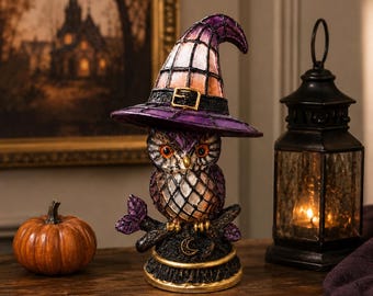 May include: A decorative owl figurine wearing a purple and orange witch hat with a gold buckle. The owl has orange eyes and sits on a dark branch base featuring a small crescent moon emblem. The piece has a textured, stained-glass style finish.
