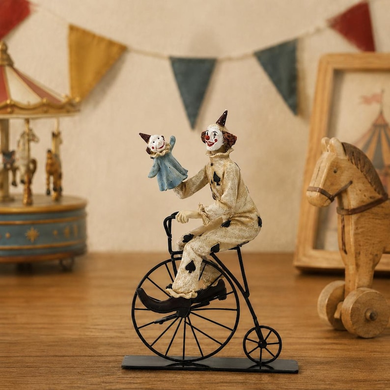 May include: A figurine of a clown wearing a cream-colored suit with black spade symbols, riding a black metal penny-farthing bicycle. The clown holds a small blue puppet with a white collar and a pointed hat, featuring a painted face with red accents.