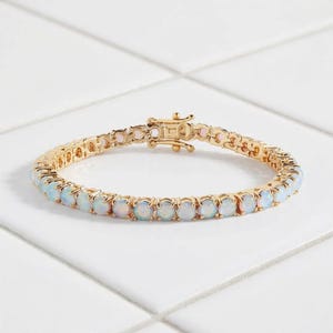 May include: A gold-tone tennis bracelet featuring a continuous row of round, iridescent white opals set in metal prongs. The bracelet includes a secure, rectangular box clasp mechanism at the top, connecting the two ends of the circular chain.
