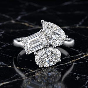 Puede incluir: Un anillo de tono plateado con tres piedras preciosas transparentes distintas en un engaste de racimo. Las piedras incluyen un corte esmeralda, una forma de pera y un corte ovalado, montadas sobre una banda de metal pulido con acabado brillante.