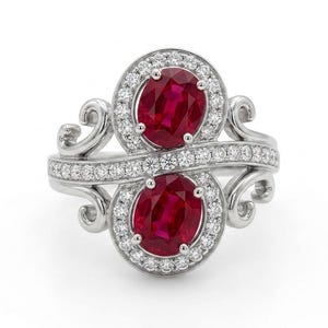 May include: A silver-toned ring featuring two oval-cut deep red rubies, each surrounded by a halo of small clear stones. The band is accented with a central row of clear stones and decorative scrollwork on both sides of the central setting.