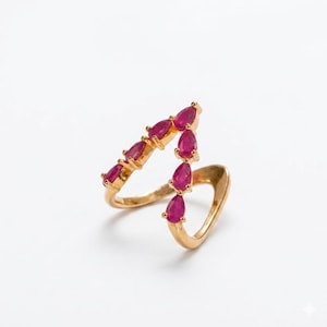 May include: A gold ring with a series of teardrop-shaped, bright red gemstones. The ring's design is asymmetrical, with the gemstones arranged along a curved line. The ring is set against a white background, highlighting its elegant design.