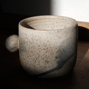 Puede incluir: Una taza de cerámica moteada con un asa redondeada. La taza es de color blanquecino con motas marrones y un diseño de remolino gris azulado oscuro. La taza está sobre una superficie de madera e iluminada por la luz solar.