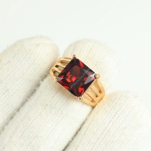 Puede incluir: Un anillo de oro con una gran piedra preciosa roja oscura de talla cuadrada. El anillo tiene un diseño clásico con una banda que tiene una serie de barras verticales a cada lado de la piedra preciosa. El anillo está sobre un fondo blanco.