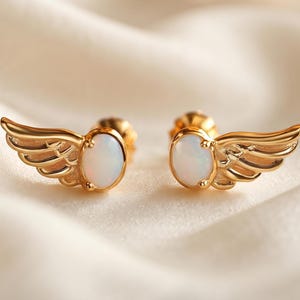 Puede incluir: Un par de pendientes de botón en forma de ala, cada uno con una piedra de ópalo blanco iridiscente de forma ovalada en el centro. Las alas tienen grabados detallados de plumas curvas y un acabado en oro pulido, con cierres visibles detrás.