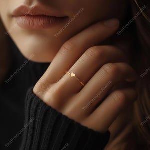 May include: A delicate gold ring featuring a small heart-shaped charm. The ring is thin and minimalist, with a polished finish. The heart charm is centered on the band. The ring is shown on a finger.