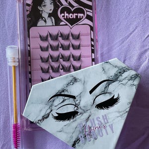 Könnte beinhalten: Ein Set aus falschen Wimpern und Zubehör. Das Set enthält eine transparente Kunststoffbox mit dem Wort "charm" und einem Bild einer Frau, eine rautenförmige Box mit den Worten "Plush Mama's Beauty" und eine Mascarabürste.