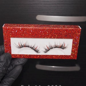 Può includere: Una scatola rossa ricoperta di glitter con una finestra trasparente che mostra un paio di ciglia finte. Le ciglia sono nere con punte rosse. Il testo "Feb 11, 2026" è visibile sotto.