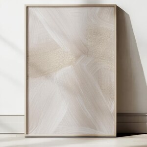 Può includere: Una stampa artistica astratta incorniciata con una tavolozza di colori neutri. L'opera d'arte presenta un design beige e bianco con motivi simili a pennellate. La cornice è di colore legno chiaro e la stampa è appoggiata a un muro bianco.