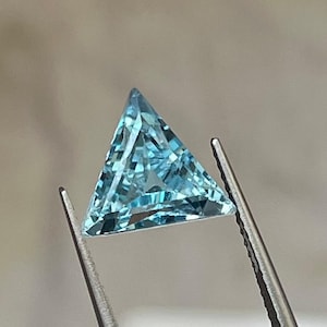 May include: A triangular, light blue gemstone held by tweezers. The gemstone has a faceted surface, reflecting light. The gem is a vibrant, clear blue color, and the shape is a perfect equilateral triangle. The tweezers are silver.